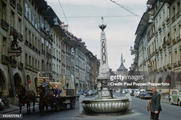 Pedestrians and a horse drawn cart pass the Kreuzgassbrunnen fountain in the middle of Kramgasse street in the centre of the city of Bern in central...