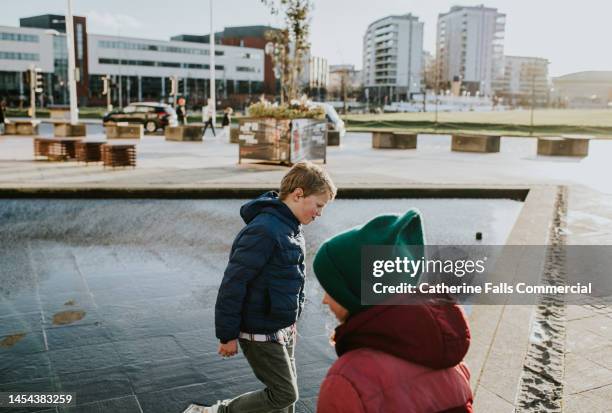 two young children explore a city, walking on a paved area beside a fountain - belfast stock pictures, royalty-free photos & images
