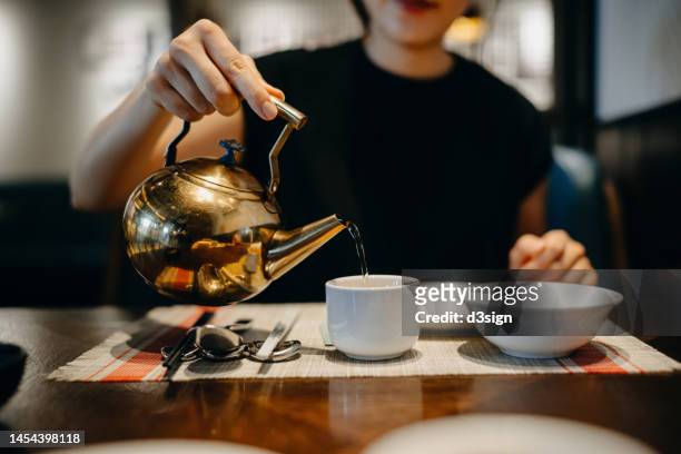 cropped shot, mid-section of young asian woman sitting in a restaurant, pouring traditional chinese tea in a cup while having meal. traditional chinese culture. yumcha, eating out lifestyle - hong kong food imagens e fotografias de stock