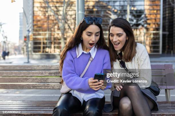 amazed young adult women looking at mobile phone screen sitting outdoors. two millennial girls using smartphone device with opened mouth showing surprise - good news bad news stock pictures, royalty-free photos & images