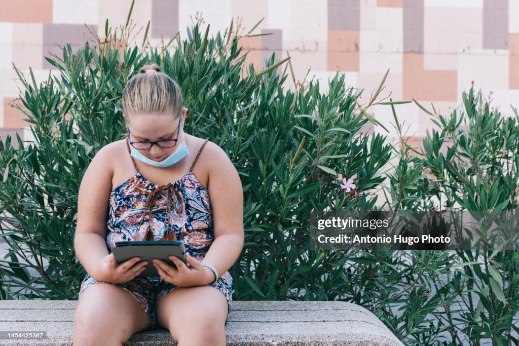Girl with glasses playing with a tablet. Sitting on a park bench.