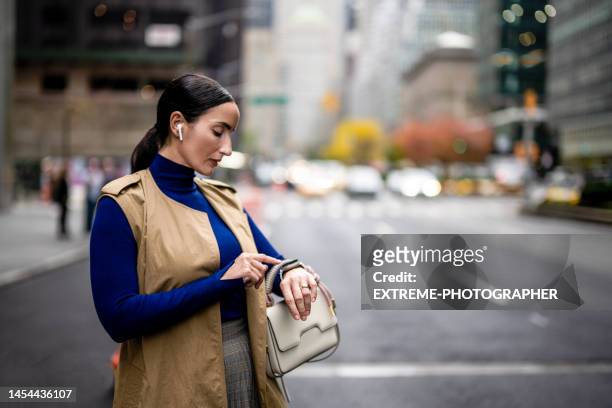 donna d'affari vista per strada di new york mentre usa il suo orologio intelligente e le cuffie wireless - sleeveless coat foto e immagini stock