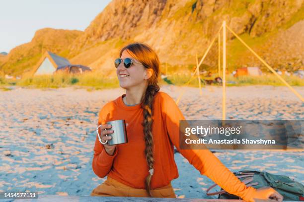 glückliche frau, die während der mitternachtssonne auf den lofoten eine tasse kaffee am strand genießt - midnight sun norway stock-fotos und bilder