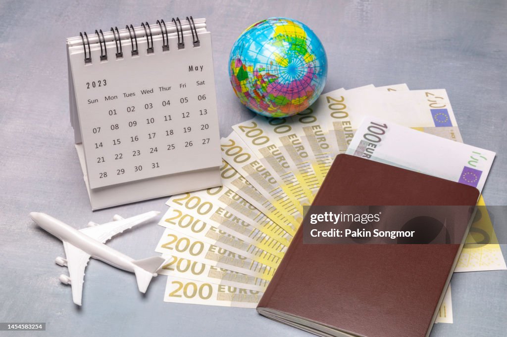 Calendar Desk 2023: May is the month for the organizer to plan and set a deadline with an airplane model, euro banknote, and passport book against an old wooden table background. travel concept.