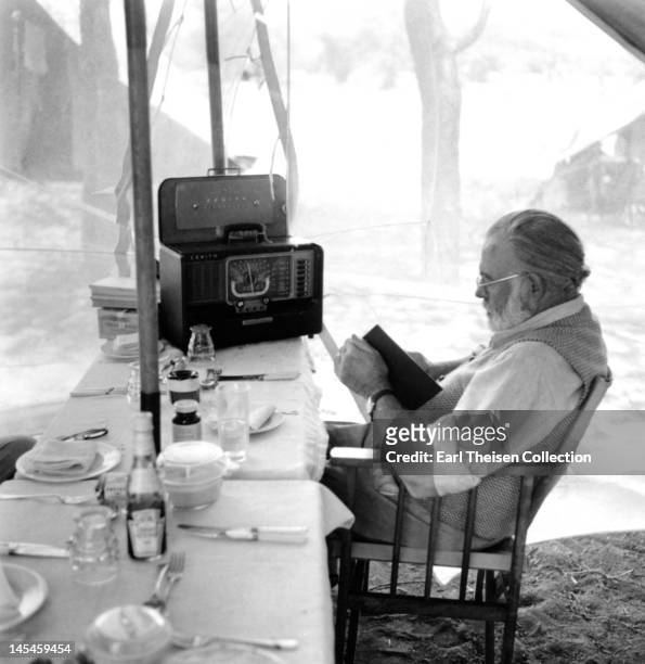 Author Ernest Hemingway reads and listens to the radio at the dining table while on a big game hunt in September 1952 in Kenya.