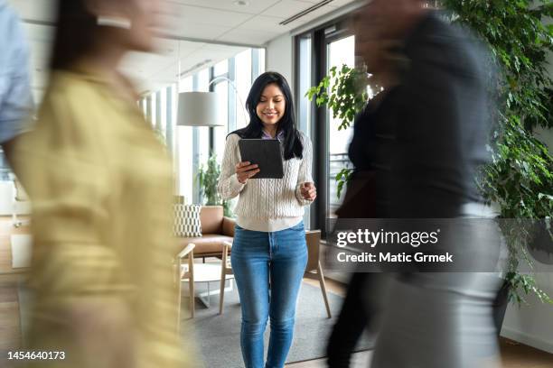 businesswoman holding digital tablet and colleagues walking by - blurred office imagens e fotografias de stock