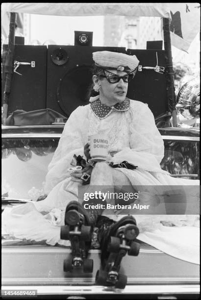 View of Rollerena Fairy Godmother, dressed in a hat, rhinestone eyeglasses, a dress, and roller skates, holding a Dunkin Donuts mug and riding on the...