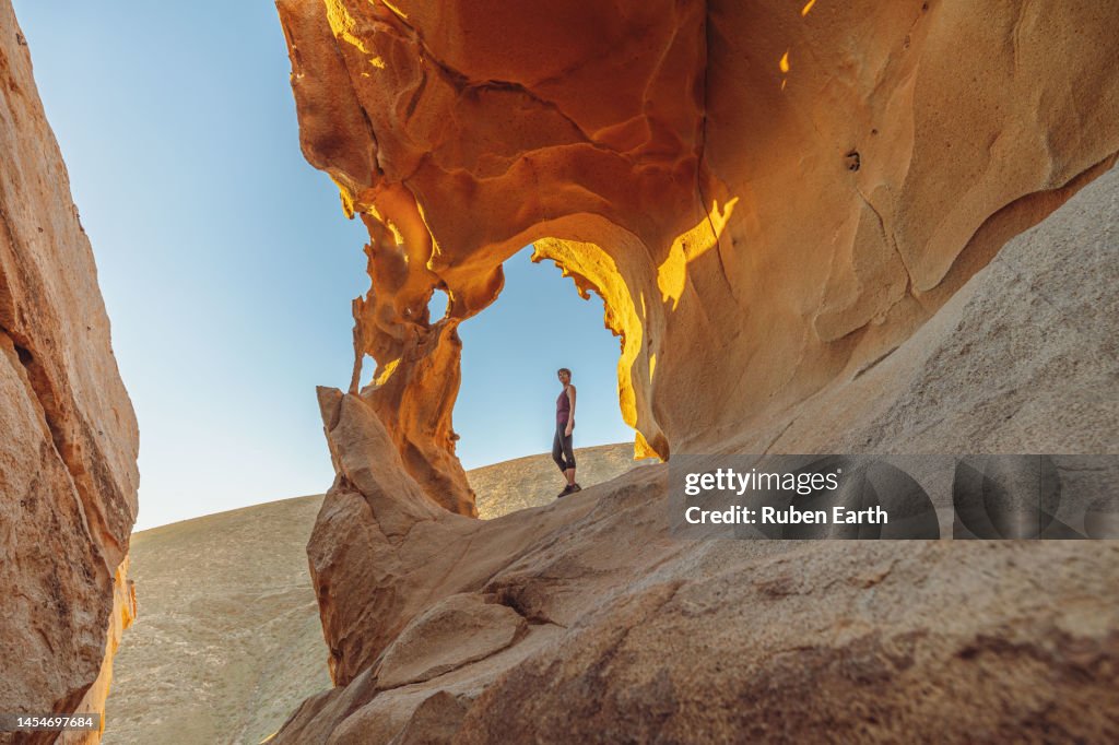 Young woman under a natural rock arch in the Fuerteventura desert at sunset