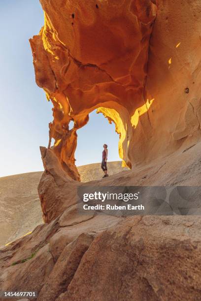 young sportswoman looking up to a volcanic rock arch in the fuerteventura desert at sunset - 33 arches stock pictures, royalty-free photos & images