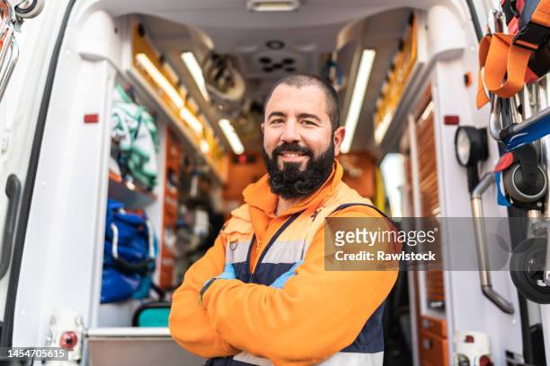portrait of a smiling young male paramedic with his arms crossed next to an ambulance - pianificazione di emergenza foto e immagini stock
