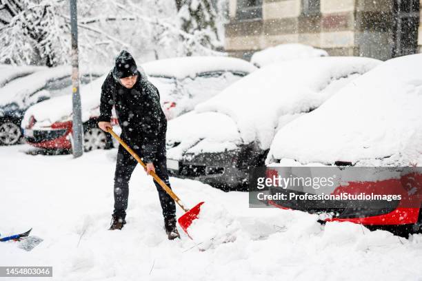 young man is cleaning a snow with shovel in front of a car. - winter snow shovel stock pictures, royalty-free photos & images