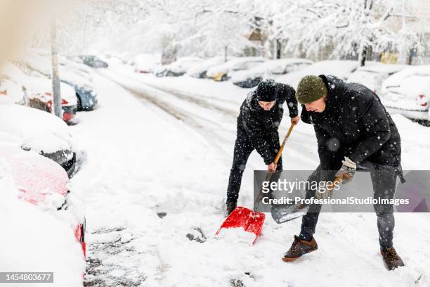 dos jóvenes están limpiando una nieve con una pala delante de un coche. - clima polar fotografías e imágenes de stock