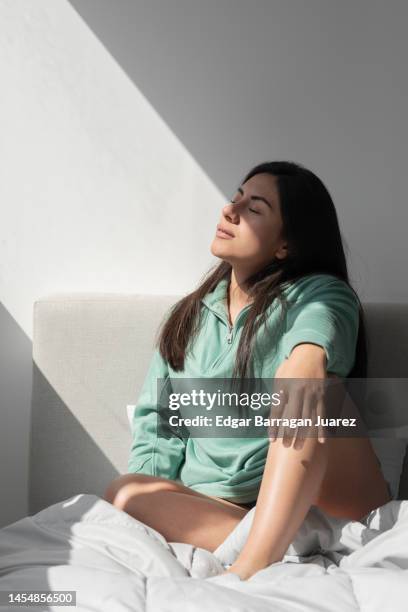 young woman sitting on her bed relaxed, while a ray of sun enters through the window, one day in the morning - alluring fitness stock pictures, royalty-free photos & images