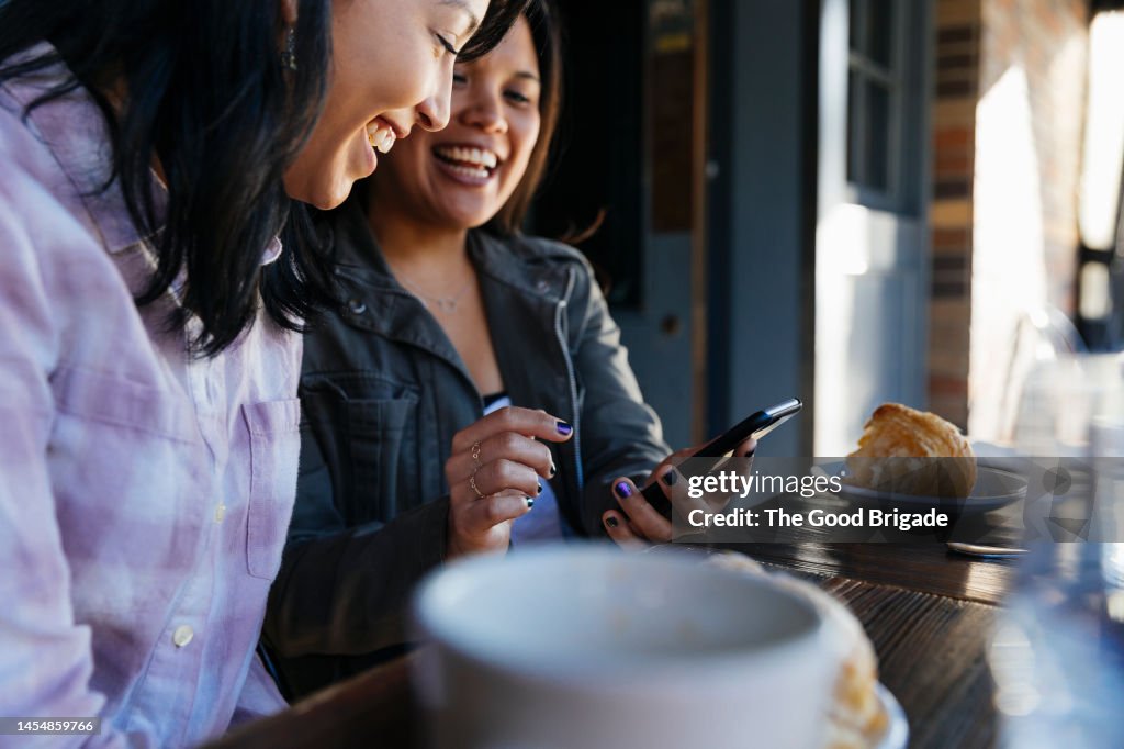 Happy woman sharing smart phone with female friend while having food in sidewalk cafe