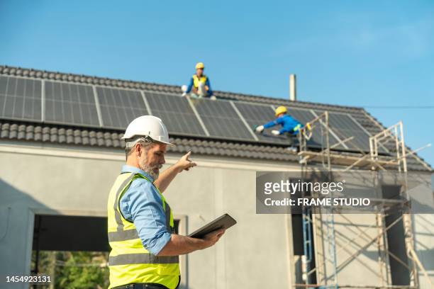 senior white european engineers directing asian workers working on installing solar panels on the roof of a house. which is a new generation of houses that reduce energy consumption and reduce the cost of electricity in the house - blank frame stockfoto's en -beelden