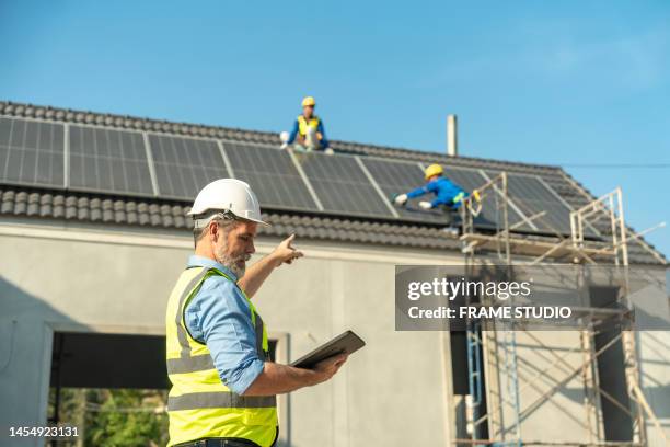 senior white european engineers directing asian workers working on installing solar panels on the roof of a house. which is a new generation of houses that reduce energy consumption and reduce the cost of electricity in the house - new generation work stock pictures, royalty-free photos & images