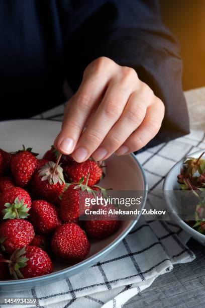 red ripe juicy delicious strawberries in a bowl or ceramic plate on a wooden background or table. a woman or a girl has a large berry in her hands, she eats it. the concept of vegetarian, vegan and raw food food and diet. - big country breakfast stock-fotos und bilder