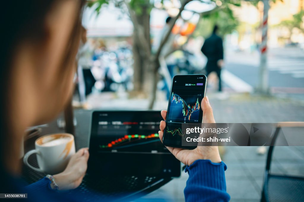 Business Woman hand using smart phone with cafe shop. Stock market charts on phone and laptop screen. Checking financial market.