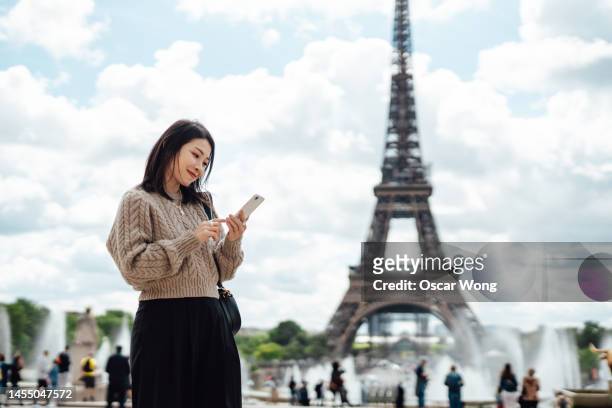 young asian woman using smartphone standing against eiffel tower in paris - tourism in paris stock pictures, royalty-free photos & images