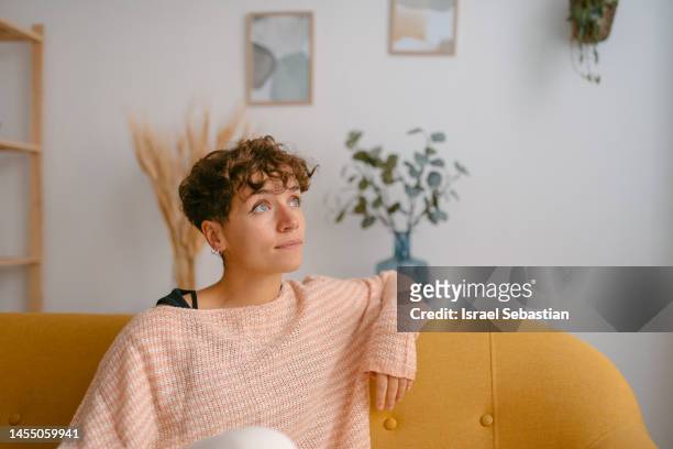 portrait of a young woman with short curly blonde hair and blue eyes, sitting on a yellow sofa, in a pensive attitude looking into the distance. - bright blue eyes stock pictures, royalty-free photos & images