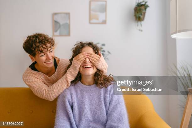 front view of a smiling young woman standing behind a sofa covering with her hands the eyes of her girlfriend to surprise her. - hands covering eyes stock pictures, royalty-free photos & images