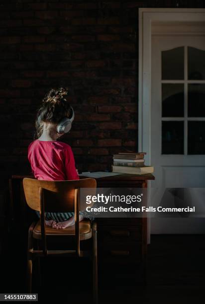 a child sits with her back to us, she is wearing headphones and studying at a wooden desk - ear plug ear protectors stock pictures, royalty-free photos & images