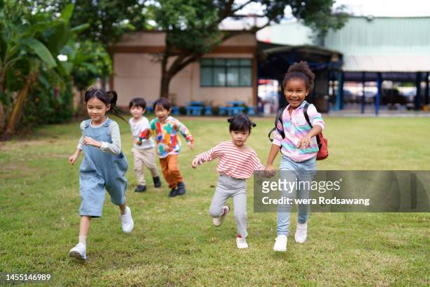 cheerful multiethnic elementary children running together on lawn of school - plat thai stock pictures, royalty-free photos & images