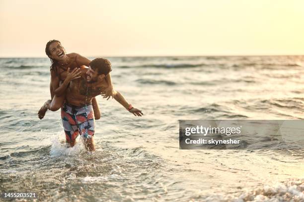 cheerful couple having fun while piggybacking in sea at sunset. - couple sunset beach stock pictures, royalty-free photos & images