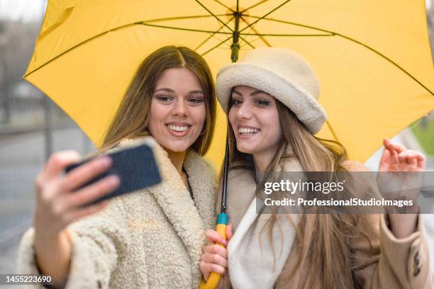 two attractive young women are walking in the city streets during a rainy day and taking a selfie photo. - rain model stockfoto's en -beelden