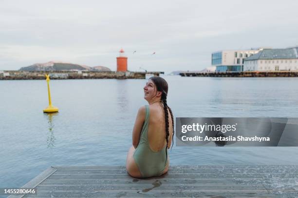rear view of female in swimsuit sitting on the pier looking at red lighthouse in norwa - winter swimming stock pictures, royalty-free photos & images