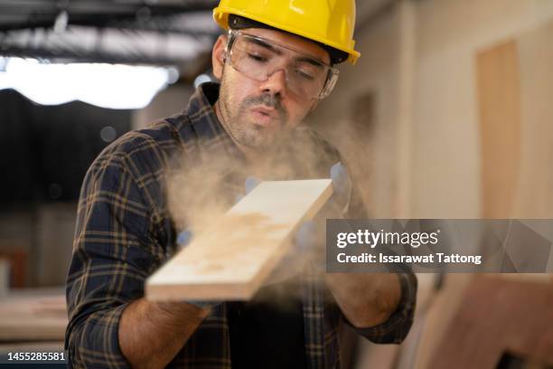 looking great. cropped shot of a focused young male carpenter blowing dust off of a piece of wood after sanding it inside of his workshop. - handyman stockfoto's en -beelden