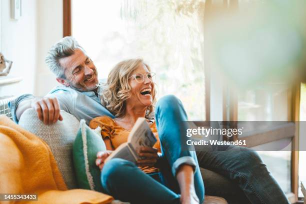 cheerful mature couple sitting on sofa at home - tous types de couple photos et images de collection