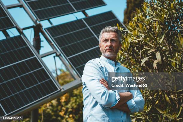 thoughtful man with arms crossed in front of solar panels on sunny day - solar panel array front view stock pictures, royalty-free photos & images