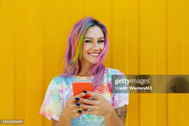happy young woman holding mobile phone in front of yellow wall - multi coloured nails stock pictures, royalty-free photos & images