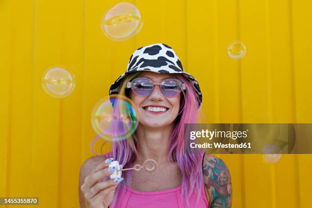 happy woman holding bubble wand in front of yellow wall - bubbles happy stockfoto's en -beelden