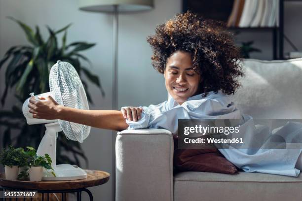 happy young woman cooling off using electric fan lying on sofa at home - ventilateur photos et images de collection