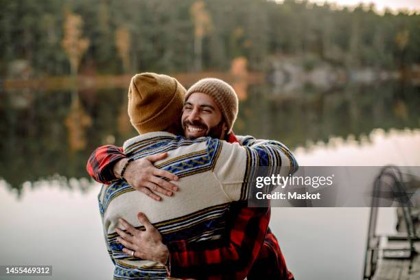 rear view of man embracing male friend near lake - bromance stock-fotos und bilder
