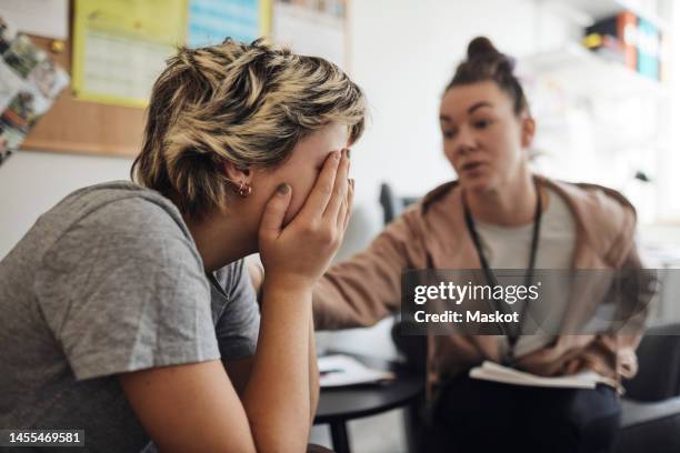 non-binary counselor consoling female student covering face with hand in school office - schoolbegeleider stockfoto's en -beelden