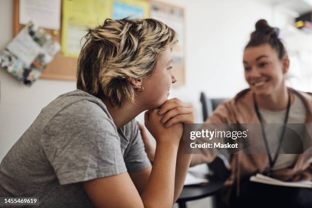 smiling mental health professional looking at female student sitting with hand on chin in school office - psicoterapeuta foto e immagini stock