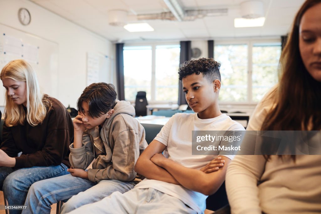 Thoughtful teenage boy sitting with arms crossed amidst friends in classroom during group therapy
