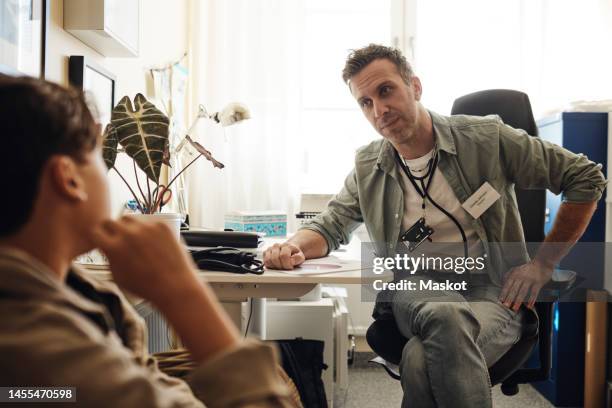 school nurse looking at teenage boy talking while sitting in office - male medical professional stock pictures, royalty-free photos & images