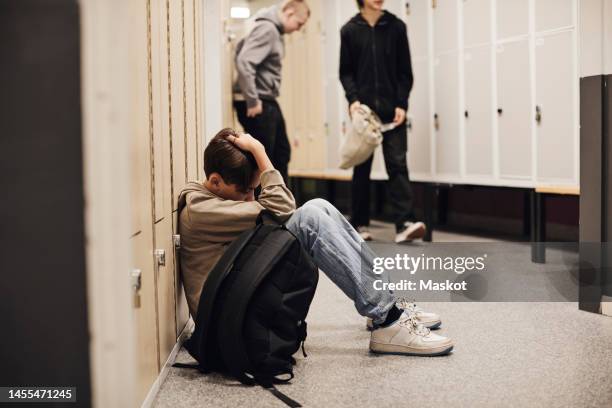 full length of depressed boy sitting with backpack in school corridor - boy sad - fotografias e filmes do acervo