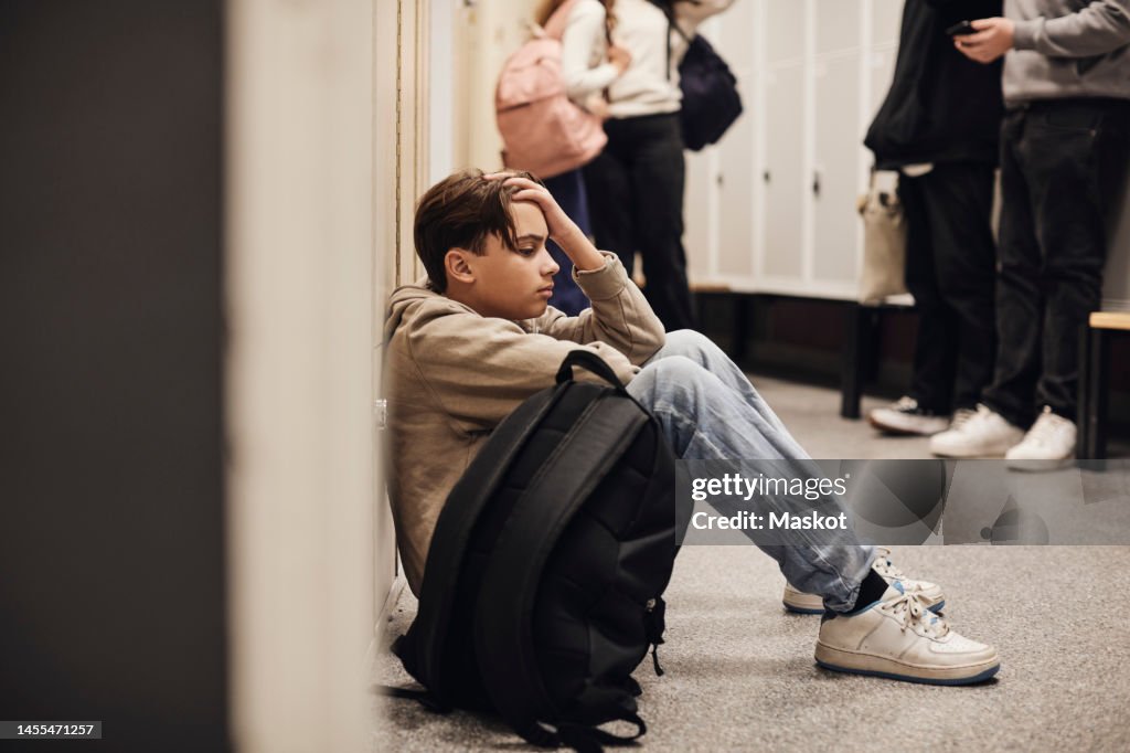 Side view of sad teenage boy sitting with backpack in school corridor