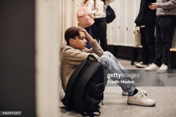 side view of sad teenage boy sitting with backpack in school corridor - mobbning bildbanksfoton och bilder