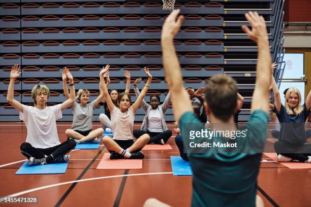 rear view of male coach with arms raised teaching yoga to students in sports court - yoga teen stock pictures, royalty-free photos & images