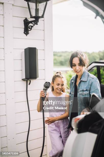 smiling daughter with mother charging electric car outside house - car home stock-fotos und bilder