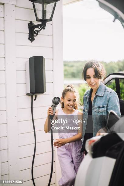 smiling daughter with mother charging electric car outside house - electric car home stock pictures, royalty-free photos & images