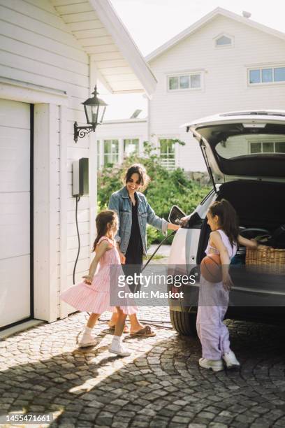 woman charging electric car while standing with daughters outside house - electric car home stock pictures, royalty-free photos & images