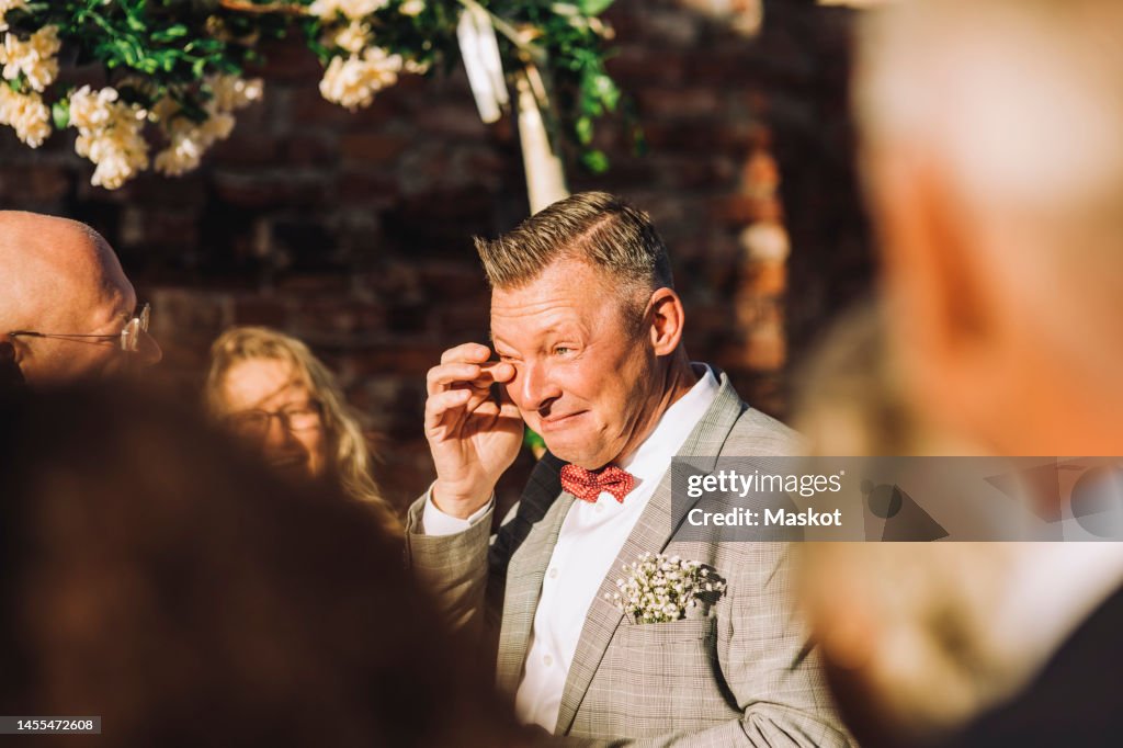 Smiling groom wiping his tears with finger amidst guests on sunny day