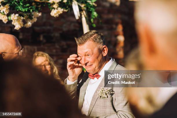 smiling groom wiping his tears with finger amidst guests on sunny day - milestone stock pictures, royalty-free photos & images